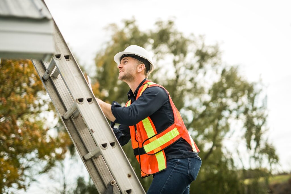 A man with hard hat standing on steps inspecting house roof