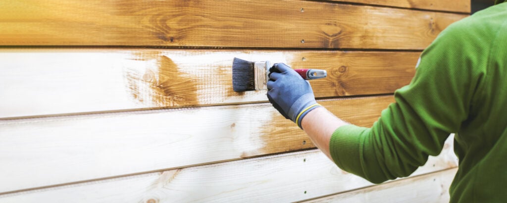 man painting wood house exterior siding with brown protective paint.